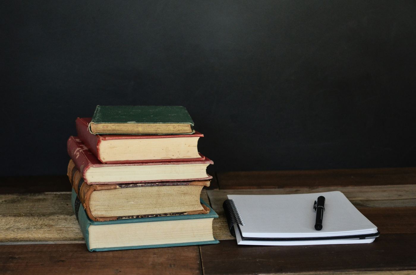 A stack of old hardback books beside an open notepad and pen