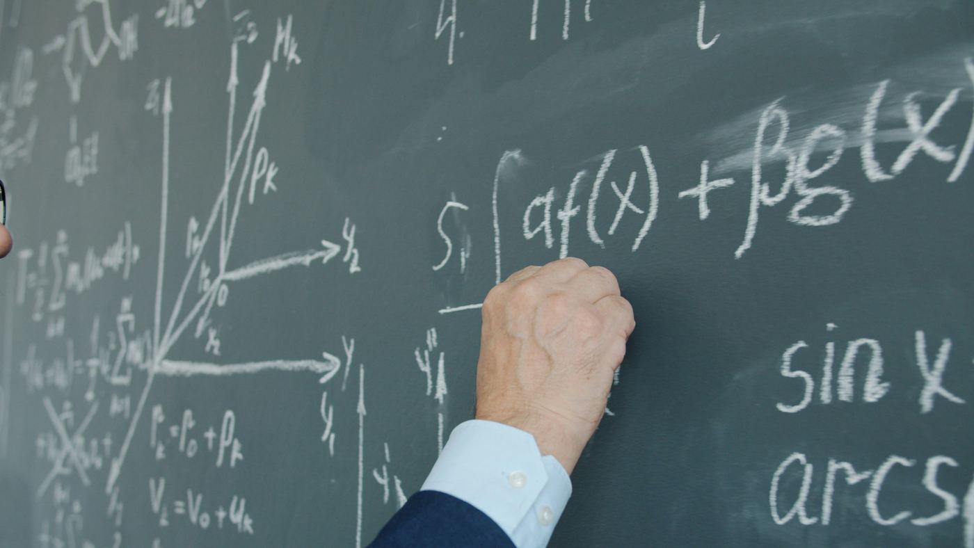 A hand writing calculus and mechanics equations on a blackboard